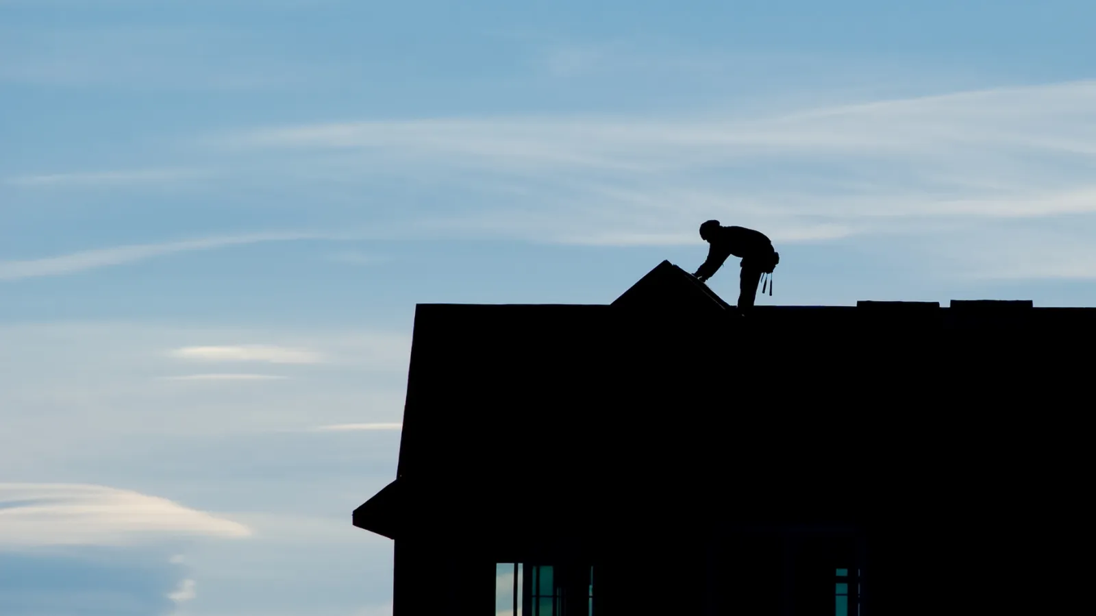 A roofer silhouetted in the distance on a house roof ridge